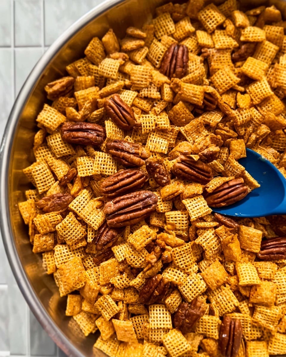 A close-up image shows a round aluminum pan filled with a snack mix. The base layer is made up of small, square, golden-brown cereal pieces with a grid texture, scattered evenly across the pan. Mixed throughout are larger, shiny, brown pecan halves that add contrast in shape and color. A blue spatula rests inside the pan, partially covered by the snack mix, hinting at recent stirring. The background features a white marbled surface. photo taken with an iphone --ar 4:5 --v 7