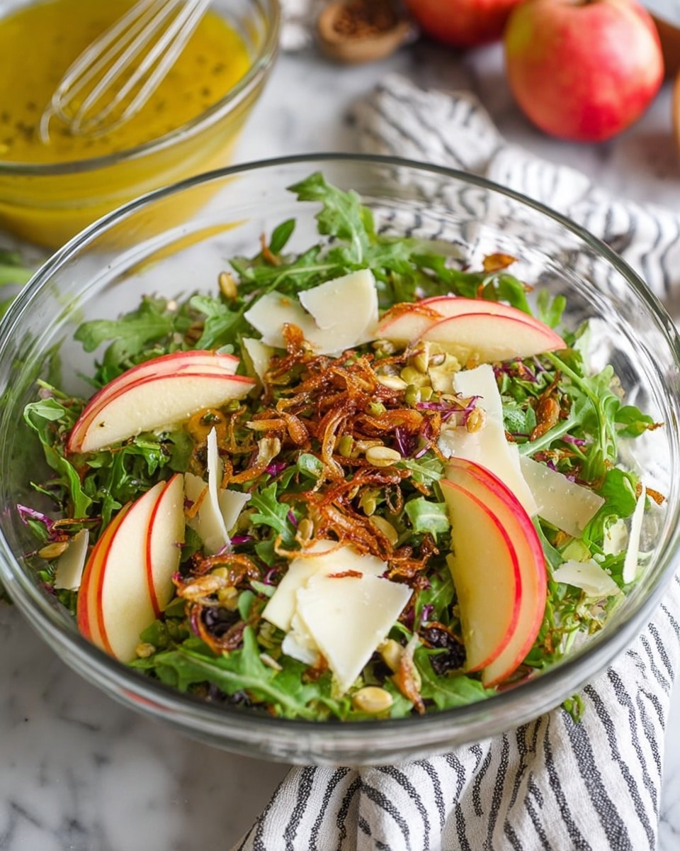 A clear glass bowl holds a fresh salad with multiple layers. The bottom layer is green arugula leaves, fresh and slightly curved. On top of this are thin slices of red apple with a shiny, smooth texture placed in small groups on opposite sides. Scattered across the salad are thin, crunchy, golden-brown fried shallots adding texture and a touch of color contrast. There are also small seeds or nuts adding detail and variety in texture throughout. Pieces of shaved white cheese with a smooth, firm texture are mixed in. The bowl is on a white marbled surface with a striped cloth nearby, and a whisk with yellow dressing is visible in the background. Photo taken with an iphone --ar 4:5 --v 7