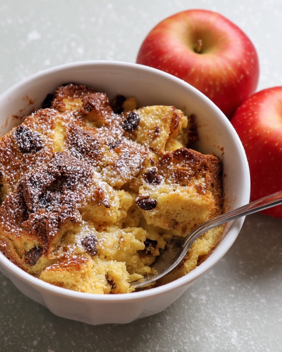 The image shows a white bowl filled with a serving of bread pudding that has a golden brown crust on top and a soft, slightly yellow interior with darker spots where raisins or chocolate chips are mixed in. The pudding is sprinkled with powdered sugar, giving it a dusted white look on the top surface. A silver spoon rests inside the bowl, partially buried in the pudding. Two red apples are placed next to the bowl on a white marbled textured surface. photo taken with an iphone --ar 4:5 --v 7