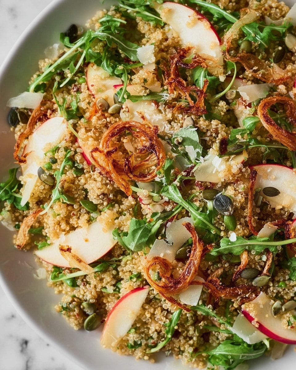 The image shows a quinoa salad with several layers closely mixed together. The base layer is made of cooked quinoa, light beige and soft in texture. Mixed evenly throughout are green arugula leaves, slightly rough and fresh-looking. Thin slices of red-skinned apple with white flesh are spread around, adding a touch of red and white smoothness. Crispy fried shallots in golden brown curls are scattered through the salad, adding a crunchy texture. Small dark green pumpkin seeds and white shaved cheese flakes are sprinkled on top, giving more color and detail to the dish. The whole salad rests on a white plate, placed on a white marbled surface. photo taken with an iphone --ar 4:5 --v 7