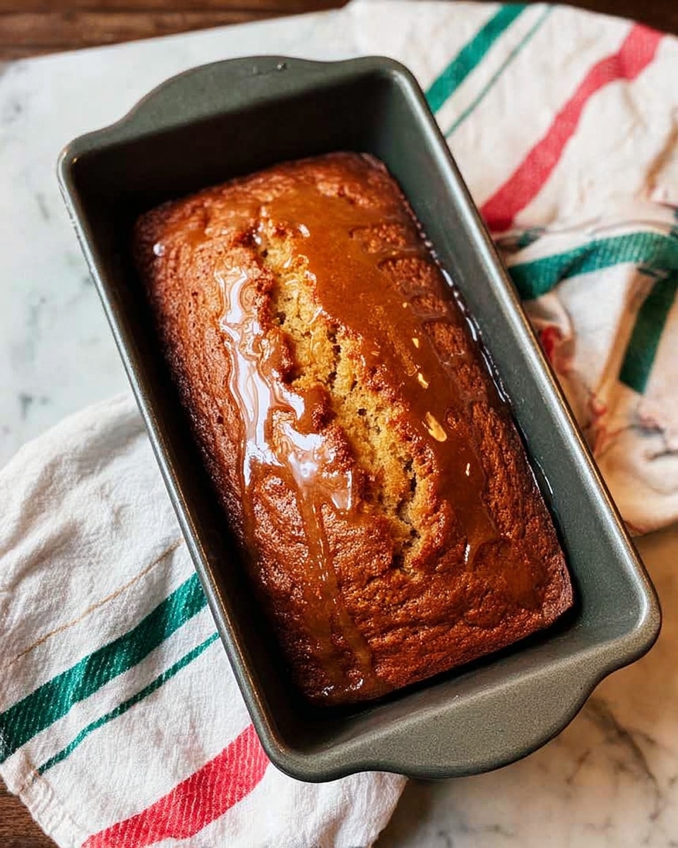 A golden-brown loaf cake sits in a dark gray metal baking pan, with a shiny glaze pooling around the edges of the cake inside the pan. The cake surface has cracks and a textured look, showing it is freshly baked. The pan rests on a white marbled surface partially covered by a crumpled white cloth with red, green, and blue stripes. The overall scene feels warm and homemade. photo taken with an iphone --ar 4:5 --v 7