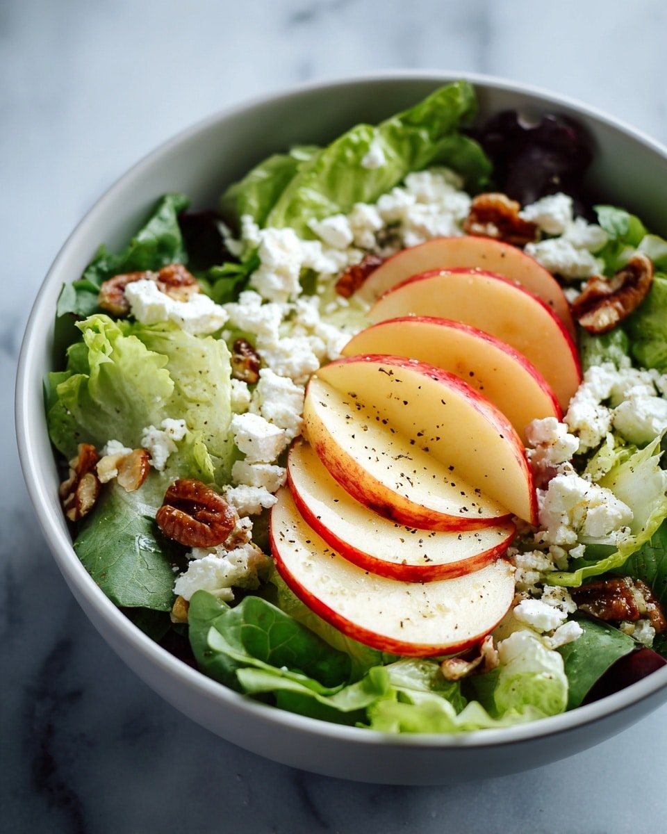 A white bowl holds a fresh salad with three main layers: the bottom layer is bright green leafy lettuce, the middle layer features thin, curved slices of red and yellow apple placed across the lettuce, and the top layer is scattered with small white chunks of cheese and brown nuts, sprinkled with black pepper and drizzled with olive oil. The bowl is set against a soft white marbled surface. photo taken with an iphone --ar 4:5 --v 7