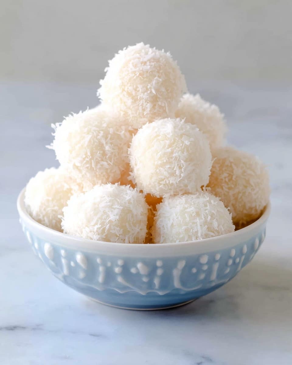 A white bowl filled with round coconut-covered balls stacked in layers, each ball completely coated in fine white shredded coconut with a soft and fluffy texture, the bowl placed on a white marbled surface. photo taken with an iphone --ar 4:5 --v 7