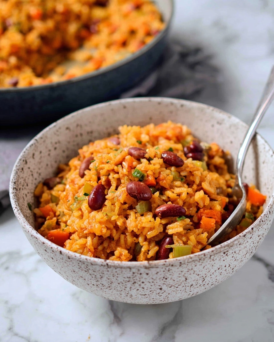 A close-up view of a bowl filled with a colorful rice dish that has two visible layers: the base layer is soft, cooked orange rice mixed with small pieces of green and orange vegetables, and the top layer is scattered reddish-brown beans. The bowl is white with a speckled grey pattern, and inside it, there's a silver spoon partially buried in the rice. In the blurred background, there is a second bowl with the same dish. The whole scene sits on a white marbled surface. Photo taken with an iphone --ar 4:5 --v 7