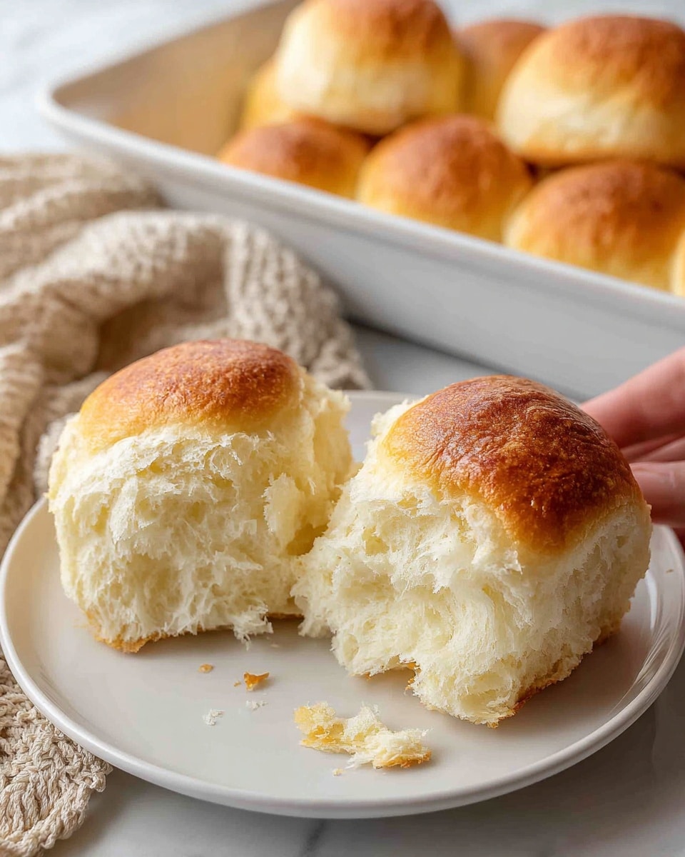 Two soft bread rolls are placed side by side on a white plate, one roll is torn open revealing a fluffy, light, and airy white inside, while the outer crust is golden brown and smooth. The torn bread shows layers of delicate, fibrous texture with small crumbs scattered near the base. In the background, there is a white baking dish filled with more golden-brown rolls, some grouped together with a similar soft texture. The scene is set on a white marbled surface with a beige woven cloth and a soft beige fabric nearby, giving a cozy feel. photo taken with an iphone --ar 4:5 --v 7