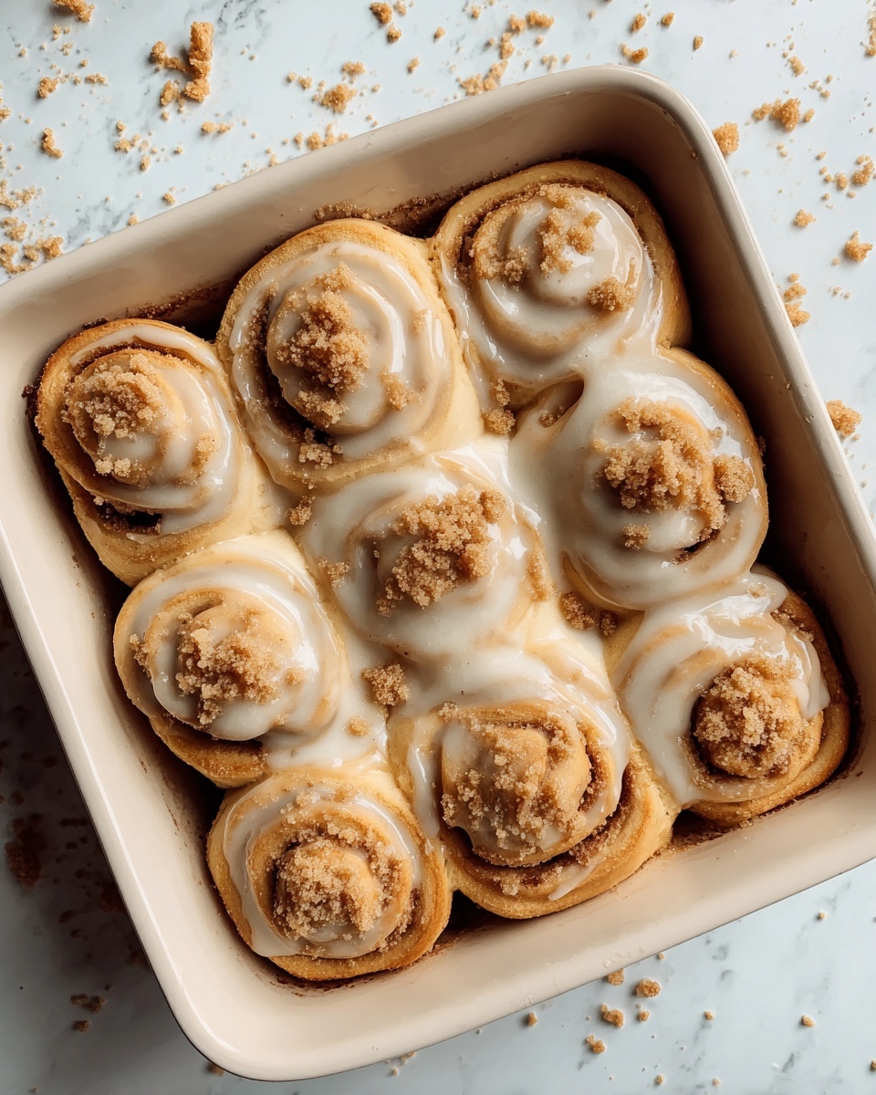 A close-up view of seven soft, golden-brown cinnamon rolls arranged tightly in a white square baking dish, each roll showing rich swirls of dough with a creamy white glaze covering the tops and flowing slightly between the layers. The glaze looks smooth and glossy, wetly dripping down the sides and pooling slightly at the bottom. Crumbly, light brown streusel bits are sprinkled generously over each roll, adding a textured contrast. The dish sits on a white marbled surface with some crumbs scattered around, enhancing the fresh-baked feel. photo taken with an iphone --ar 4:5 --v 7