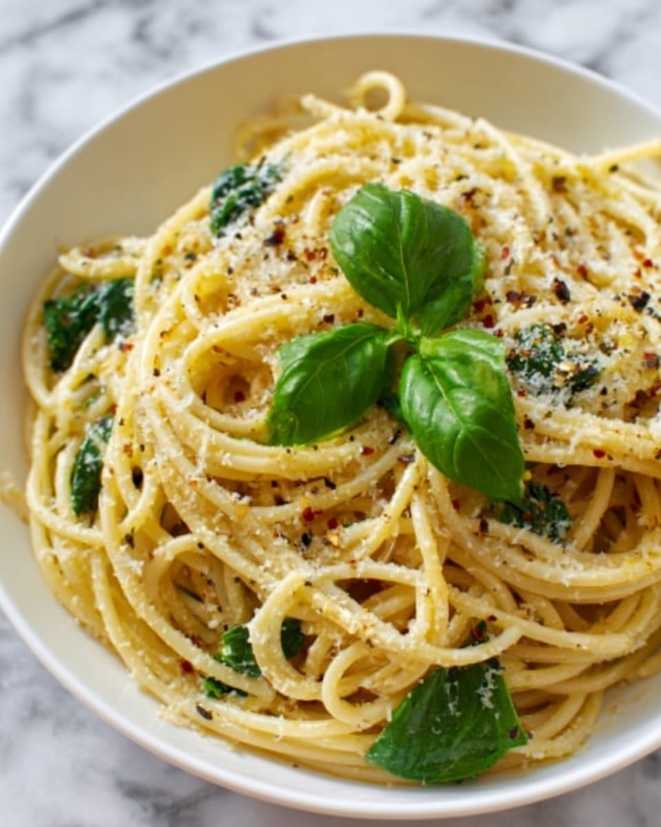 A close-up of a white bowl filled with spaghetti pasta mixed with wilted green spinach leaves, small pieces of black pepper, and grated cheese sprinkled on top. The pasta strands are lightly coated in a creamy sauce with a slightly yellow tint. A fresh basil leaf rests on the top layer as garnish. The background shows a smooth white marbled surface. photo taken with an iphone --ar 4:5 --v 7