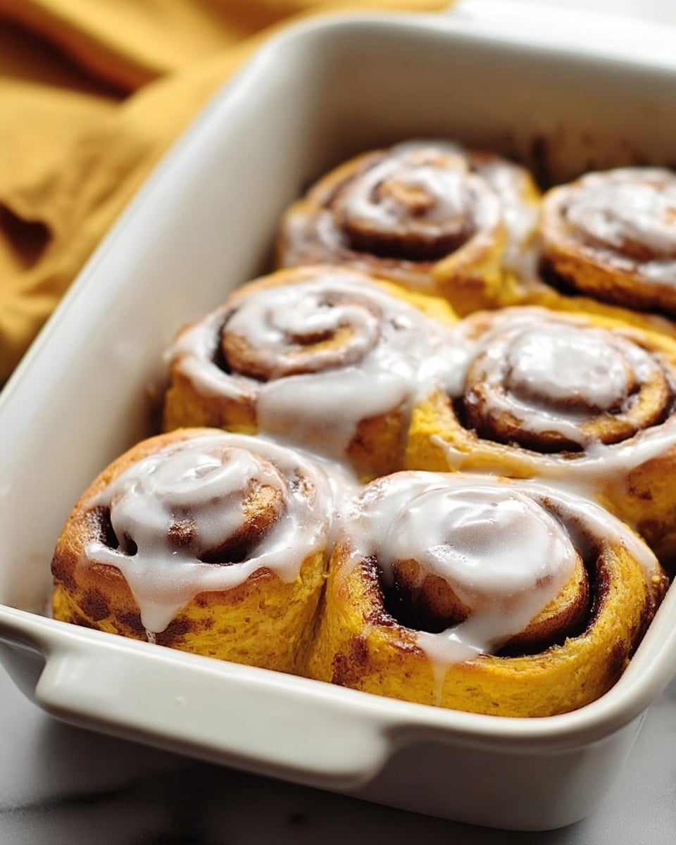 In a white rectangular baking dish, six pumpkin-colored cinnamon rolls are tightly placed in two rows of three. Each cinnamon roll has a spiral shape with visible layers showing dark brown cinnamon sugar filling inside. The rolls are topped with a glossy white glaze that lightly spreads inside the spirals and pools a little around the bottom edges. The surface below the dish has a white marbled texture, and there is a soft golden cloth blurred in the background. photo taken with an iphone --ar 4:5 --v 7