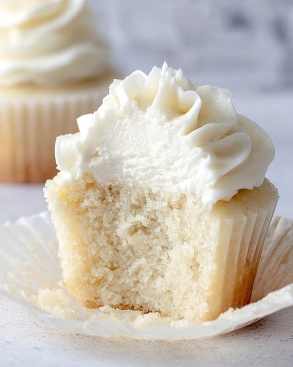 A close-up image shows a single white cupcake that is cut in half. The cupcake has two main layers: the first layer is a soft, light, fluffy white cake with a moist texture, positioned as the base. The second layer on top is a thick, creamy white frosting, swirled smoothly and slightly peaked in the center, with a soft and airy texture. The cupcake sits in a white paper liner that is spread open around it. The background has a white marbled texture, with a blurred second cupcake in the distance. Photo taken with an iphone --ar 4:5 --v 7
