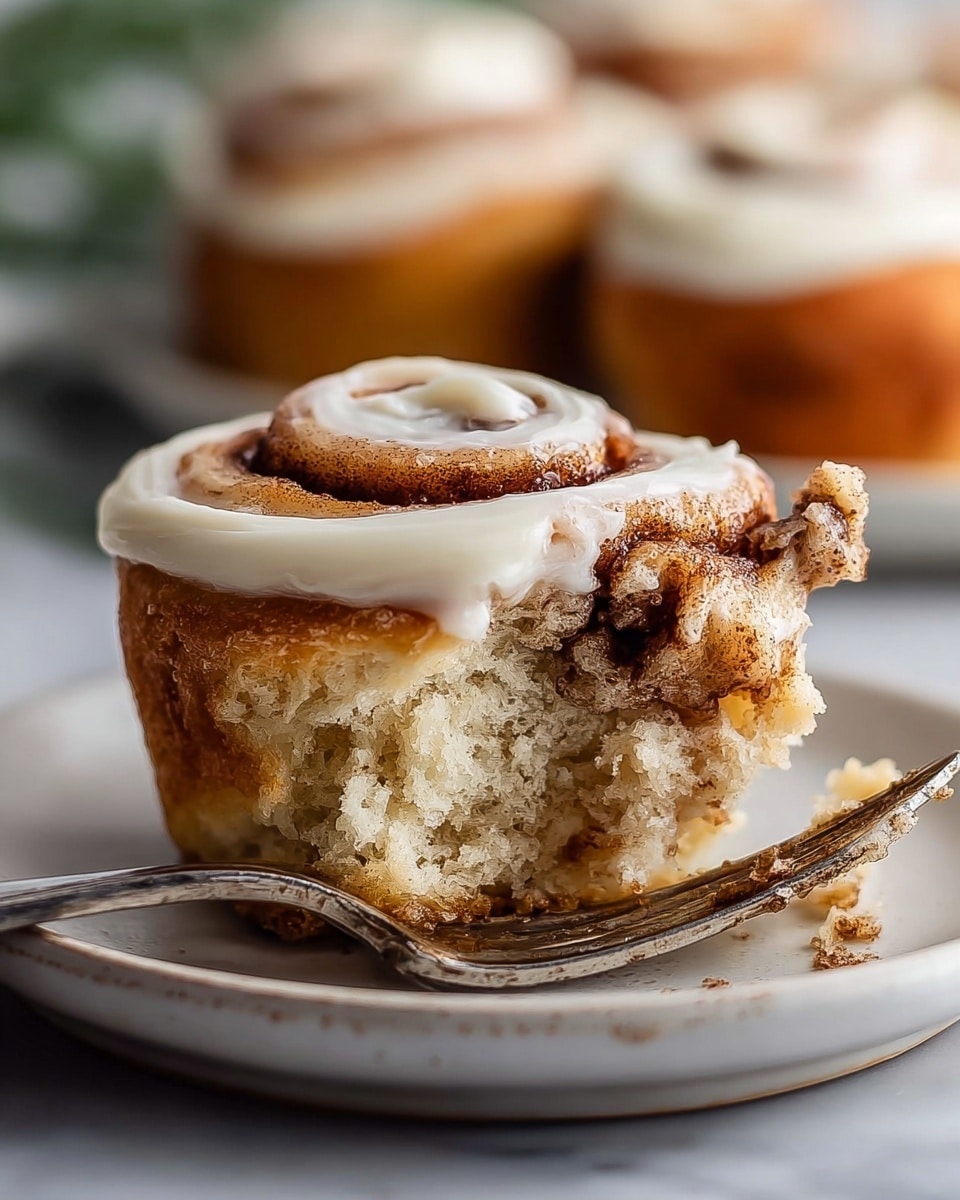 A close-up image of a cinnamon roll with three visible layers: the bottom layer is a golden-brown, slightly crispy crust, the middle layer is soft and light beige dough with a swirl of dark cinnamon filling, and the top layer is a thick swirl of creamy white frosting partially covering the cinnamon swirl. The cinnamon roll sits on a white plate with a slightly uneven edge, placed on a white marbled surface. In the foreground, a silver fork is lifting a bite of the cinnamon roll, showing the soft inside texture and cinnamon filling, with a blurred background of more cinnamon rolls. Photo taken with an iphone --ar 4:5 --v 7