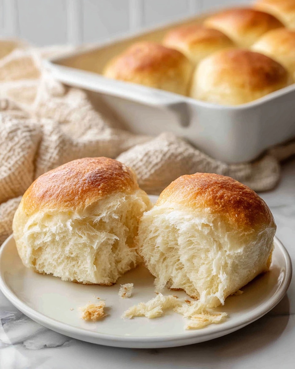 Two soft, golden brown bread rolls sit closely on a simple white plate. Each roll has a shiny, smooth top crust that fades into a light, fluffy inside showing delicate layers and a slightly torn texture from being pulled apart. The plate rests on a textured cloth with more rolls visible in a white baking dish and on the white marbled surface behind, creating a cozy, fresh-from-the-oven scene. photo taken with an iphone --ar 4:5 --v 7
