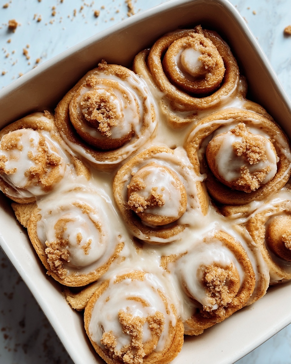 The image shows a close-up of seven cinnamon rolls in a white square baking dish, arranged tightly in two rows with four rolls in one row and three in the other, each roll having a golden brown color with visible smooth swirls of dough. A creamy white glaze is generously spread over the tops and between the rolls, creating a shiny and slightly thick layer that glistens in the light. Sprinkle of light brown sugar granules is scattered unevenly on top of each roll, adding texture and contrast. The baking dish is placed on a white marbled surface with some crumbs scattered around it. Photo taken with an iphone --ar 4:5 --v 7