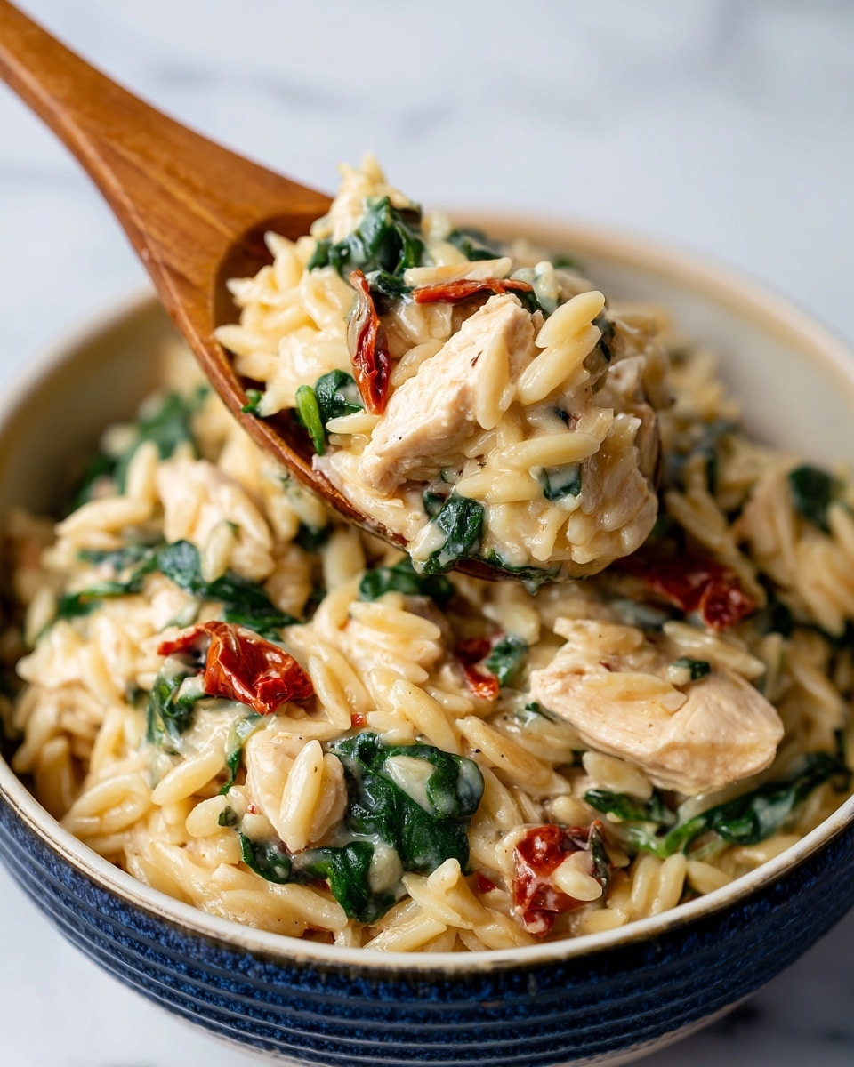The image shows a close-up of a blue bowl filled with creamy orzo pasta mixed with chunks of white chicken, wilted dark green spinach leaves, and pieces of deep red sun-dried tomatoes. A wooden spoon is scooping up a portion of the dish, highlighting the soft texture of the pasta and the tender chicken. The background features a white marbled texture surface. Photo taken with an iphone --ar 4:5 --v 7