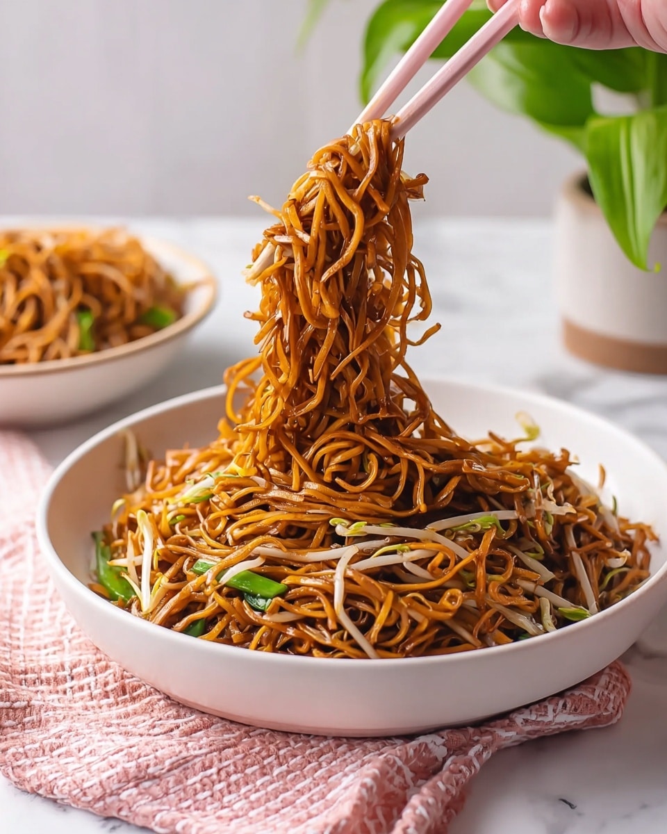 A large white bowl filled with stir-fried noodles that are brown and slightly shiny, mixed with light-colored bean sprouts and green vegetables, with a woman's hand using light pink chopsticks to lift a thick bundle of noodles above the bowl. In the background, there is another white bowl with more noodles, and a green plant in a white square pot against a white marbled surface with a pink and white checkered cloth partially visible in the foreground. photo taken with an iphone --ar 4:5 --v 7
