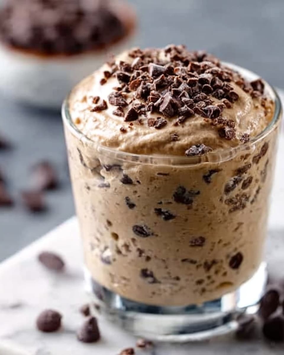 A clear glass filled with a creamy, light brown mixture dotted with small chocolate chips and sprinkled with more chocolate chips on top, showing a thick texture and rich look. The glass sits on a white marbled surface, with a blurred white bowl filled with chocolate chips in the background. Photo taken with an iphone --ar 4:5 --v 7