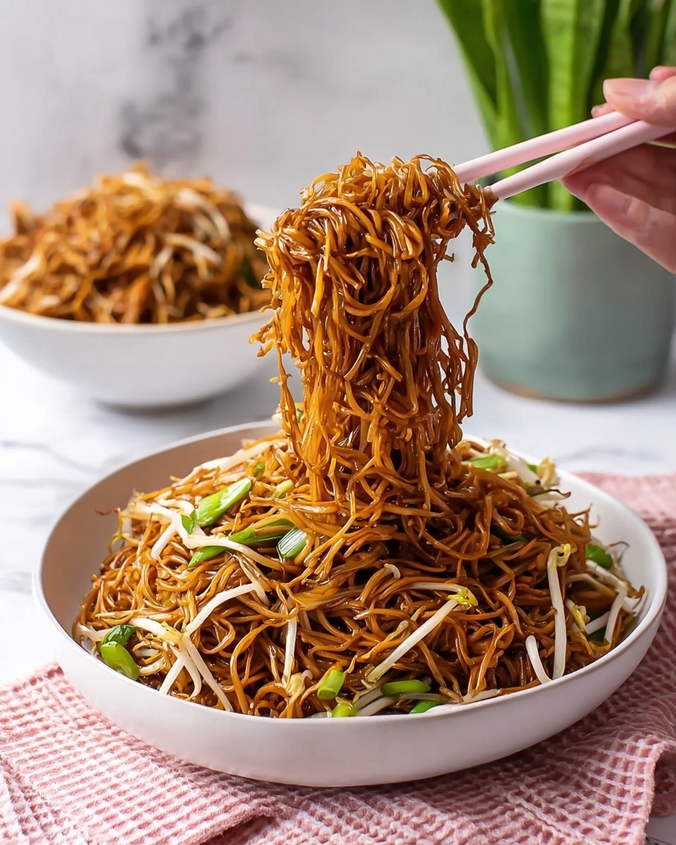 A bowl of stir-fried noodles with a glossy dark brown color fills a white bowl, mixed with thin light beige bean sprouts and small pieces of green vegetables scattered throughout. A woman's hand holding light pink chopsticks lifts a tangled clump of noodles above the bowl, showing the noodles' thin and slightly curly texture. In the background, there is a second white bowl filled with more noodles, partially blurred. The setting is on a white marbled surface with a pink and white checkered cloth at the front and a green potted plant on the right side. photo taken with an iphone --ar 4:5 --v 7