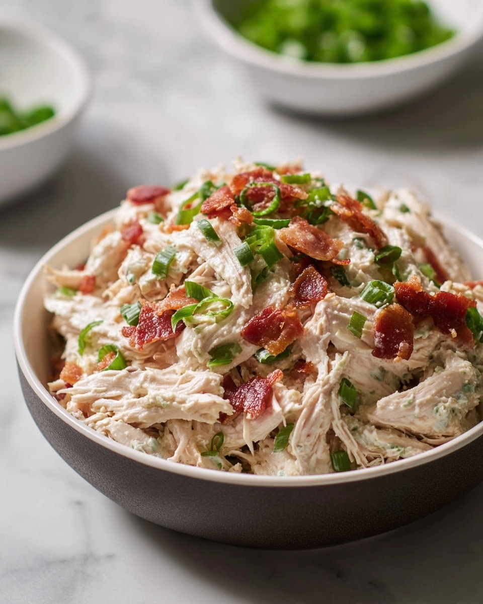 A close-up view of a bowl filled with shredded white chicken mixed with creamy dressing, topped with green chopped scallions and small crispy reddish-brown bacon pieces scattered throughout. The bowl is white on the inside and dark on the outside, sitting on a white marbled surface, with two blurred white bowls in the background, one containing green chopped vegetables. The texture of the chicken looks tender and moist, combined with the smooth creamy layer and crunchy bacon bits. photo taken with an iphone --ar 4:5 --v 7