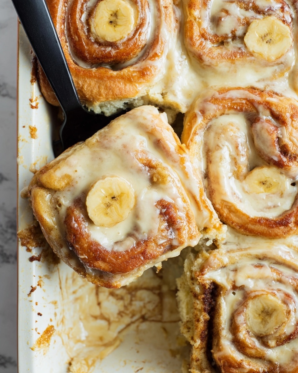 A close-up view of a baking tray holding soft cinnamon rolls covered in creamy icing. Each roll has two to three visible layers of golden-brown dough with a glossy texture from the creamy icing that fills the gaps and covers the top. Thin slices of banana sit on the surface of the rolls, adding a pale yellow contrast against the warm brown dough and light cream topping. A black spatula lifts one roll, showing its moist and fluffy inside with swirled cinnamon filling. The white baking tray has browned bits around the edges, suggesting a fresh bake. The background is a white marbled texture photo taken with an iphone --ar 4:5 --v 7