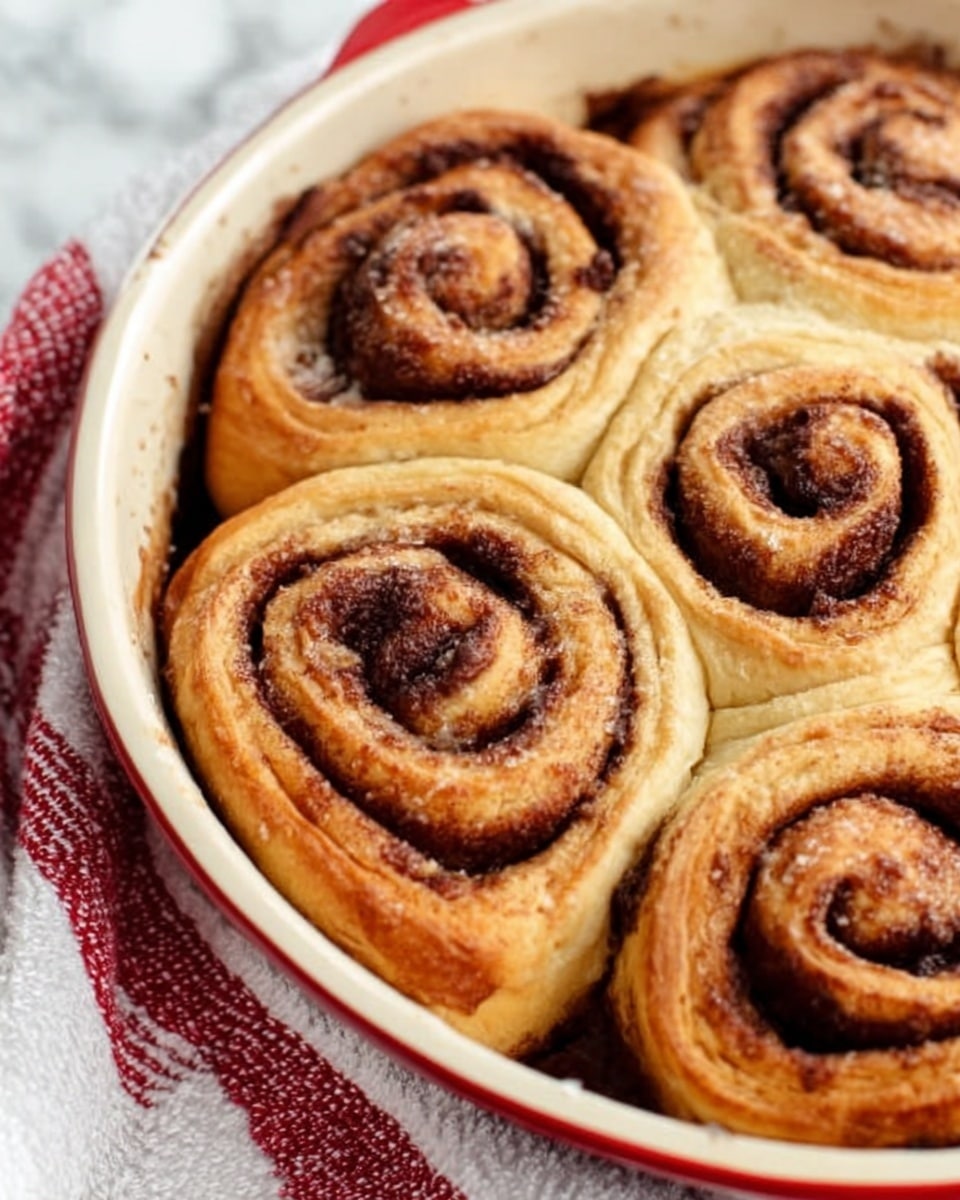 A close-up view of several freshly baked cinnamon rolls arranged closely in a white ceramic dish with a red rim. Each cinnamon roll has multiple layers of soft, golden-brown dough spiraled with dark brown cinnamon filling that looks sticky and sweet. The layers are thick and fluffy, showing a clear swirl pattern. The dish is placed on a white marbled surface with a folded white cloth with red stripes under it. The lighting highlights the warmth and texture of the rolls, making them look soft and inviting. Photo taken with an iphone --ar 4:5 --v 7