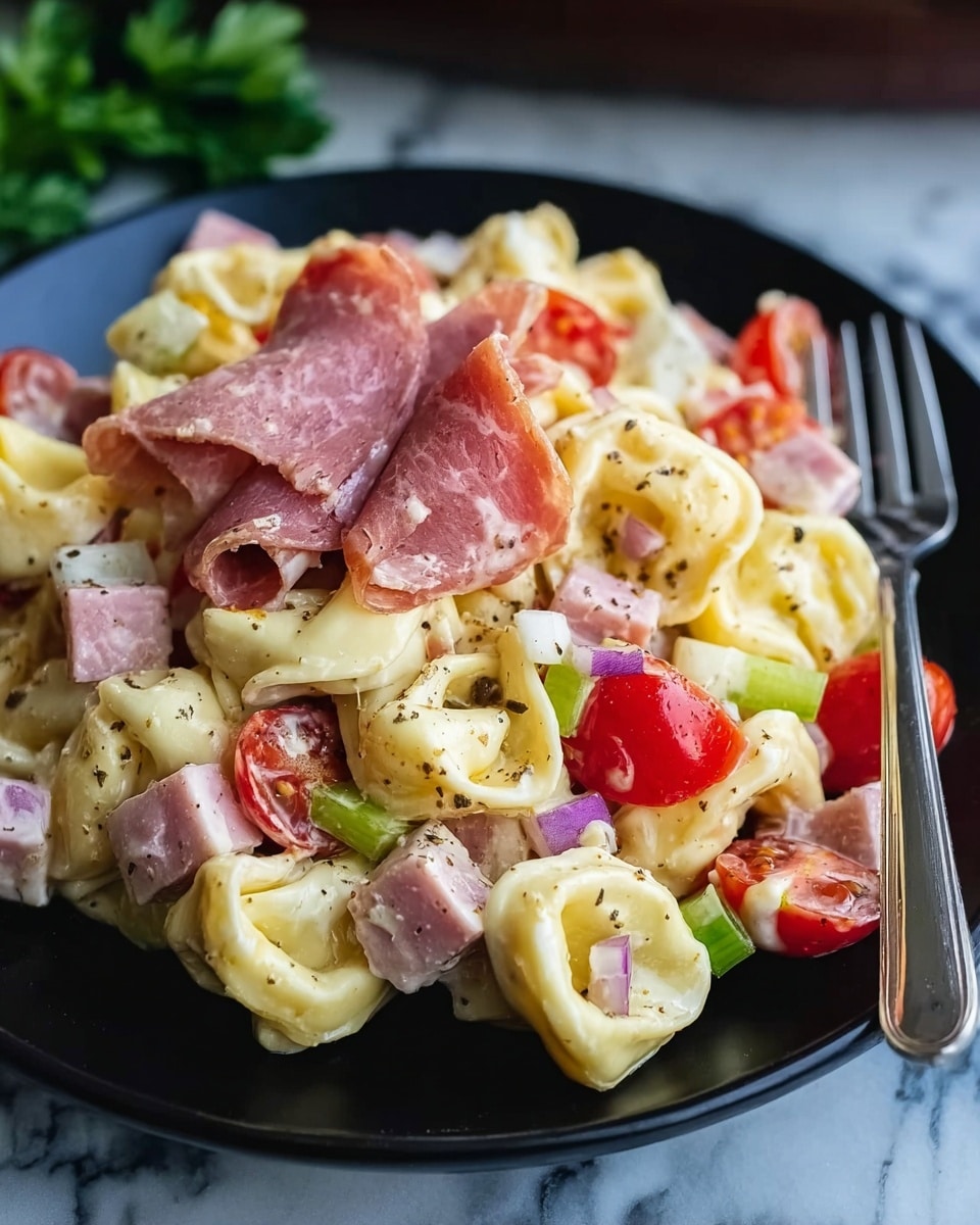 A close-up view of a black plate filled with creamy tortellini pasta layered with pale yellow cheese-filled pasta, bright red cherry tomato halves, small cubes of white cheese, light green chopped celery, thin slices of pink ham, and bits of purple onion, all mixed together and sprinkled with black pepper, placed on a white marbled surface with a fork on the right side of the plate and a blurred red tomato and green herbs in the background, photo taken with an iphone --ar 4:5 --v 7