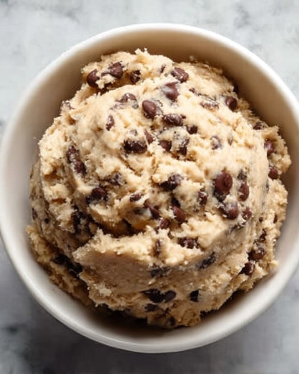 A close-up of a round scoop of cookie dough with many dark brown chocolate chips scattered throughout. The dough is light beige with a slightly rough texture, full of small lumps and chocolate chips. It sits in a white bowl with smooth edges, placed on a white marbled surface. The lighting highlights the uneven surface and the rich chocolate pieces in the dough. photo taken with an iphone --ar 4:5 --v 7