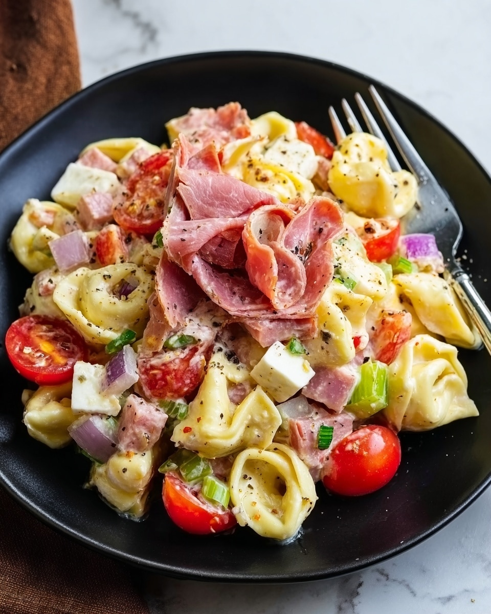 A black plate is filled with a layered tortellini pasta salad, with creamy pale yellow tortellini as the base layer, mixed with bright red cherry tomato halves scattered throughout. Cubes of light pink ham and white cheese are layered evenly, along with bits of purple onion and small pieces of green celery adding pops of color. Thin slices of pink cured meat rest on top, with visible black pepper sprinkled across the whole dish. A silver fork leans against the side on a white marbled surface background. photo taken with an iphone --ar 4:5 --v 7