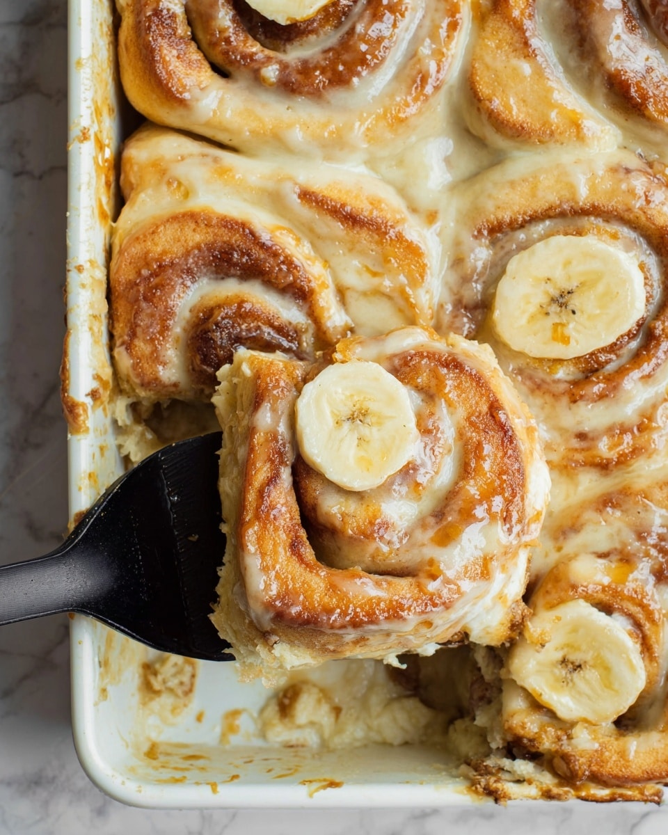 A close-up of a square cinnamon roll in a white baking tray, being lifted with a black spatula, showing a swirl of golden-brown dough with a creamy beige icing on top. Each cinnamon roll has one visible banana slice pressed into the iced surface near the center, surrounded by soft, shiny swirls with a slightly caramelized texture. The tray has some browned crumbs and spots, adding texture to the scene. The background is a white marbled texture. Photo taken with an iphone --ar 4:5 --v 7